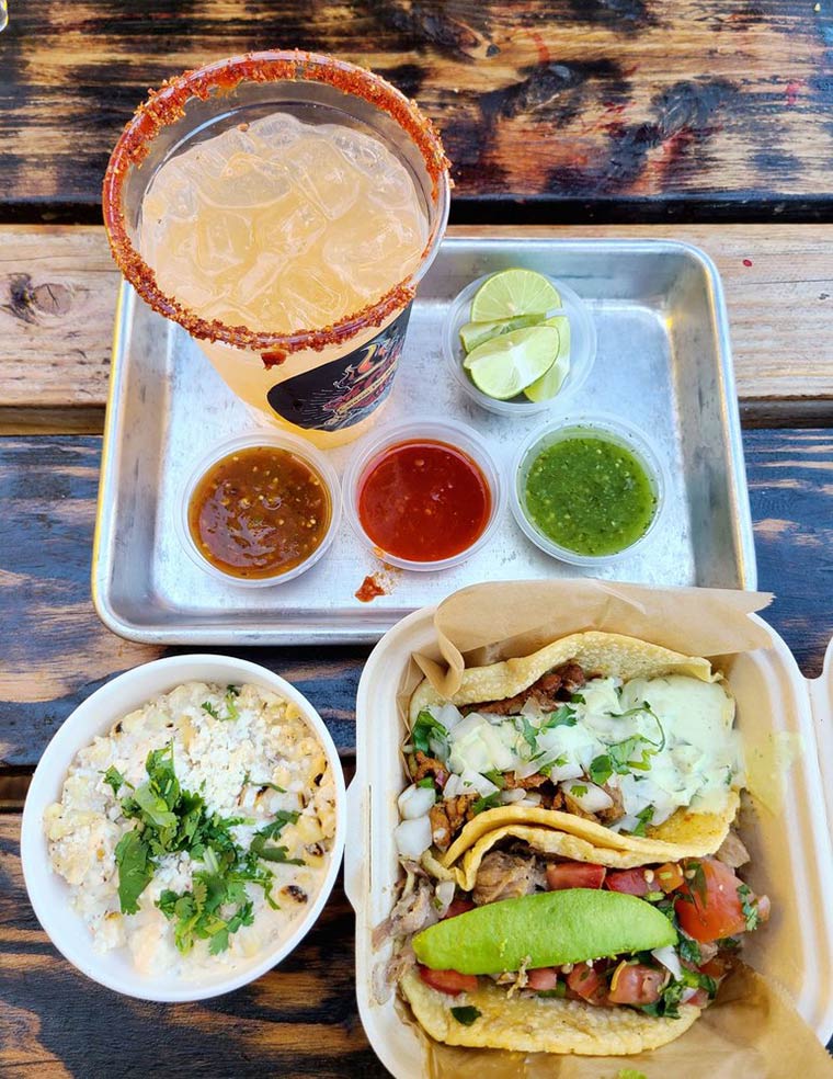 Delicious tachos served in a to-go box, accompanied by an array of flavorful salsas and a zesty spicy margarita.