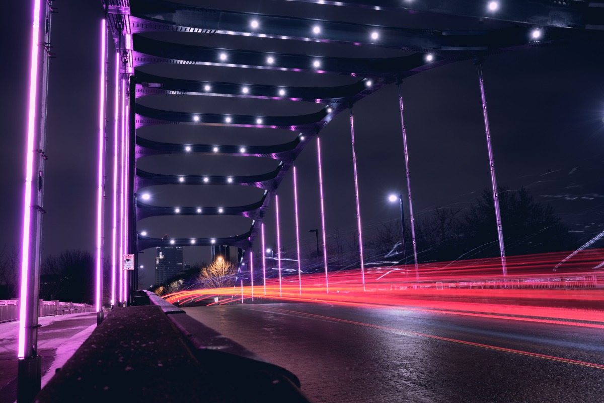 Purple LED lights on a bridge in Fort Wayne, IN lit up at night. A long exposure of car brake lights runs through the shot.