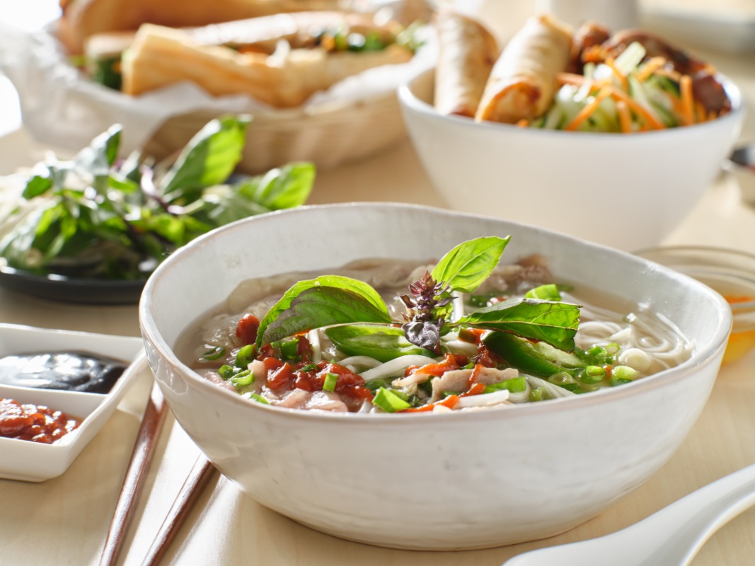 A white ceramic bowl full of Pho sits on a table among other Vietnamese style dishes in the background.