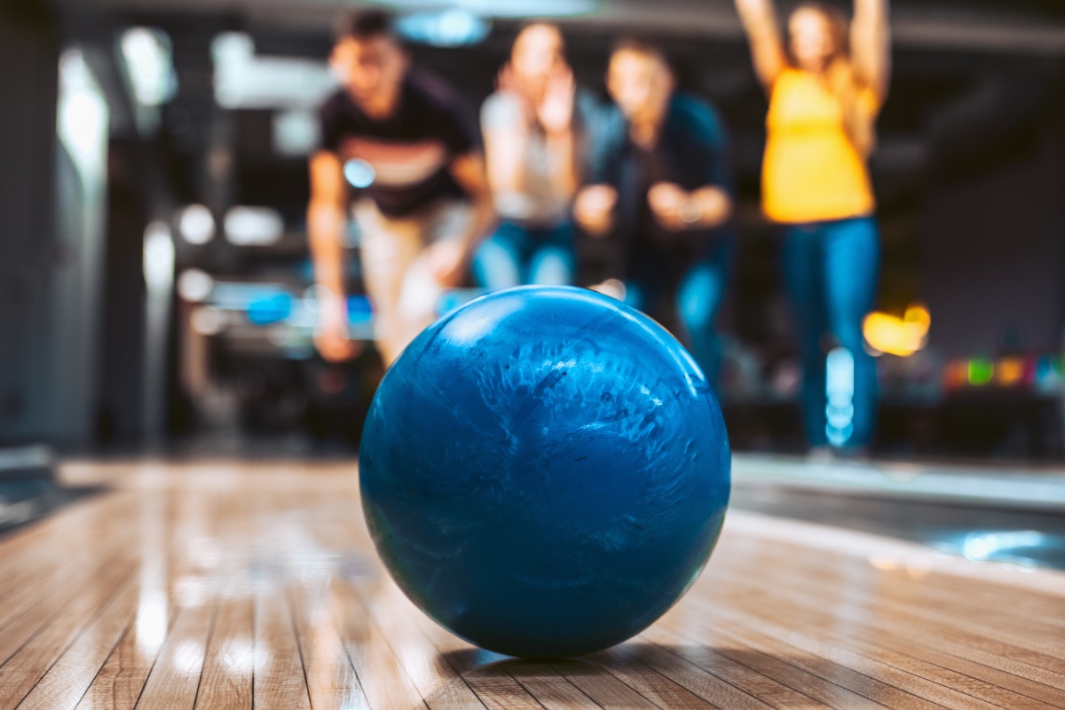 A blue bowling ball is thrown down the lane towards the camera. People are cheering in the background. 