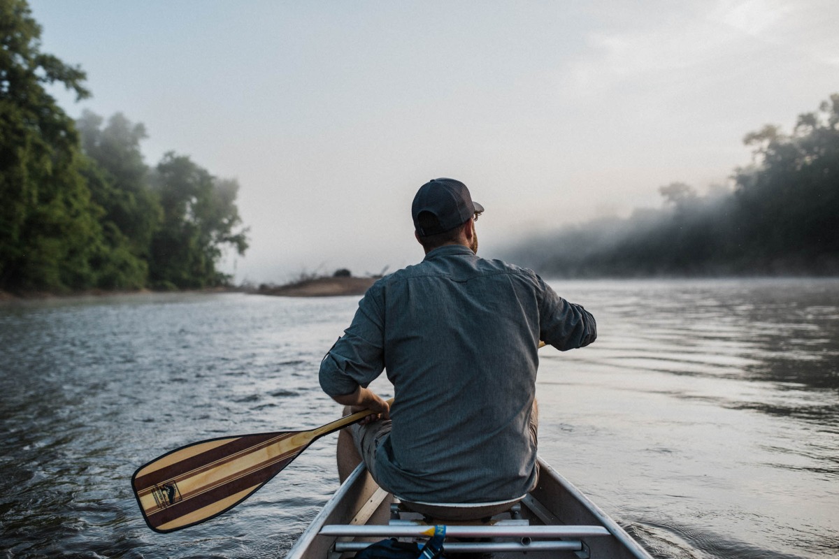 A man paddles a canoe on the water. The lighting is fresh and there is a slight mist in the sky. 