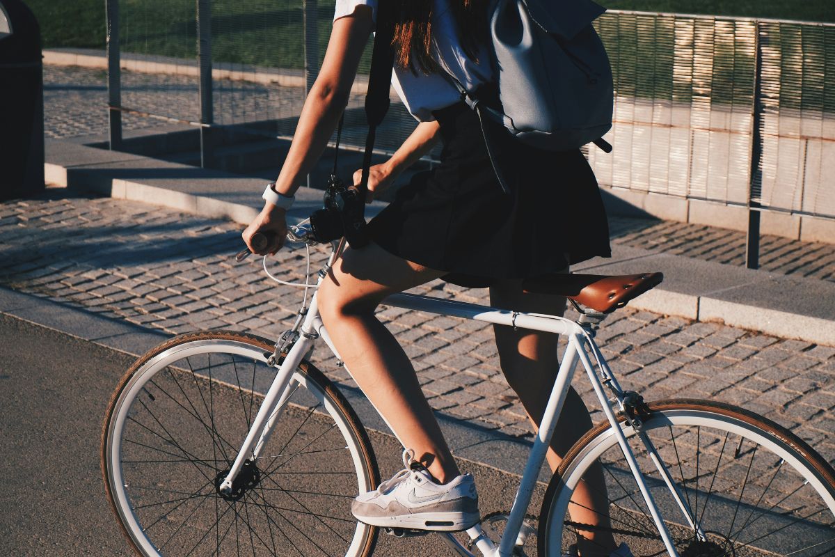 A girl in black skirt and wearing a small backpack leads a classic bike in a street in the city.