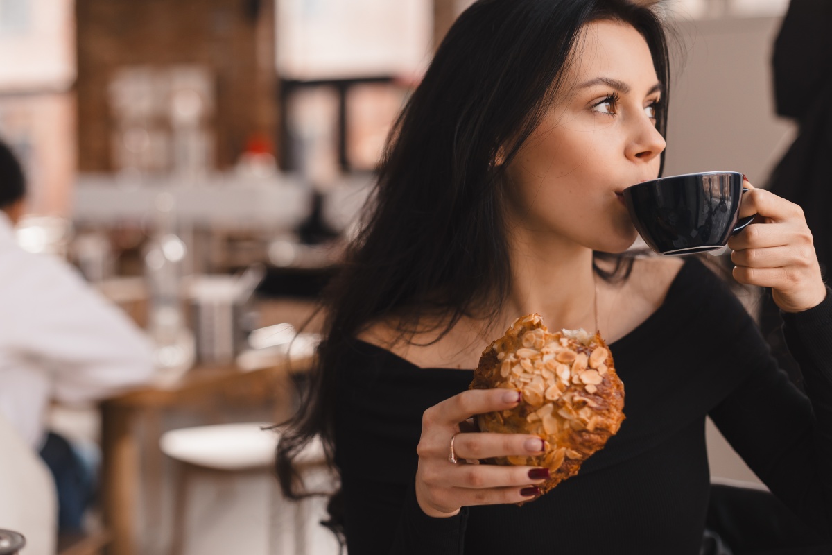 A young woman takes a sip of coffee from a black cup in a local cafe and holds a pastry in her hands. 