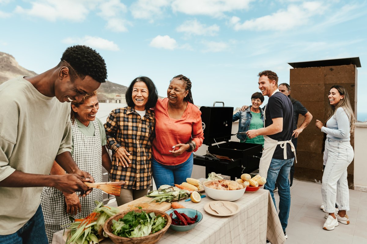 Un groupe d'amis et de membres de la famille parle et rit sur une table de style buffet installé sur un toit d'appartement aux côtés d'un grill. 